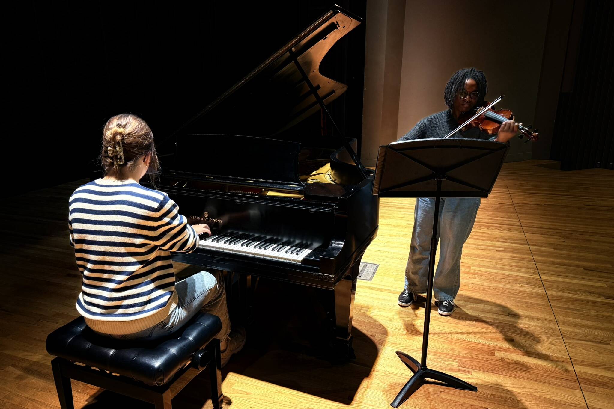 Two musicians perform on stage; one plays a grand piano while seated, wearing a striped sweater, and the other stands playing a violin, using a music stand. The stage has wooden flooring and is dimly lit.