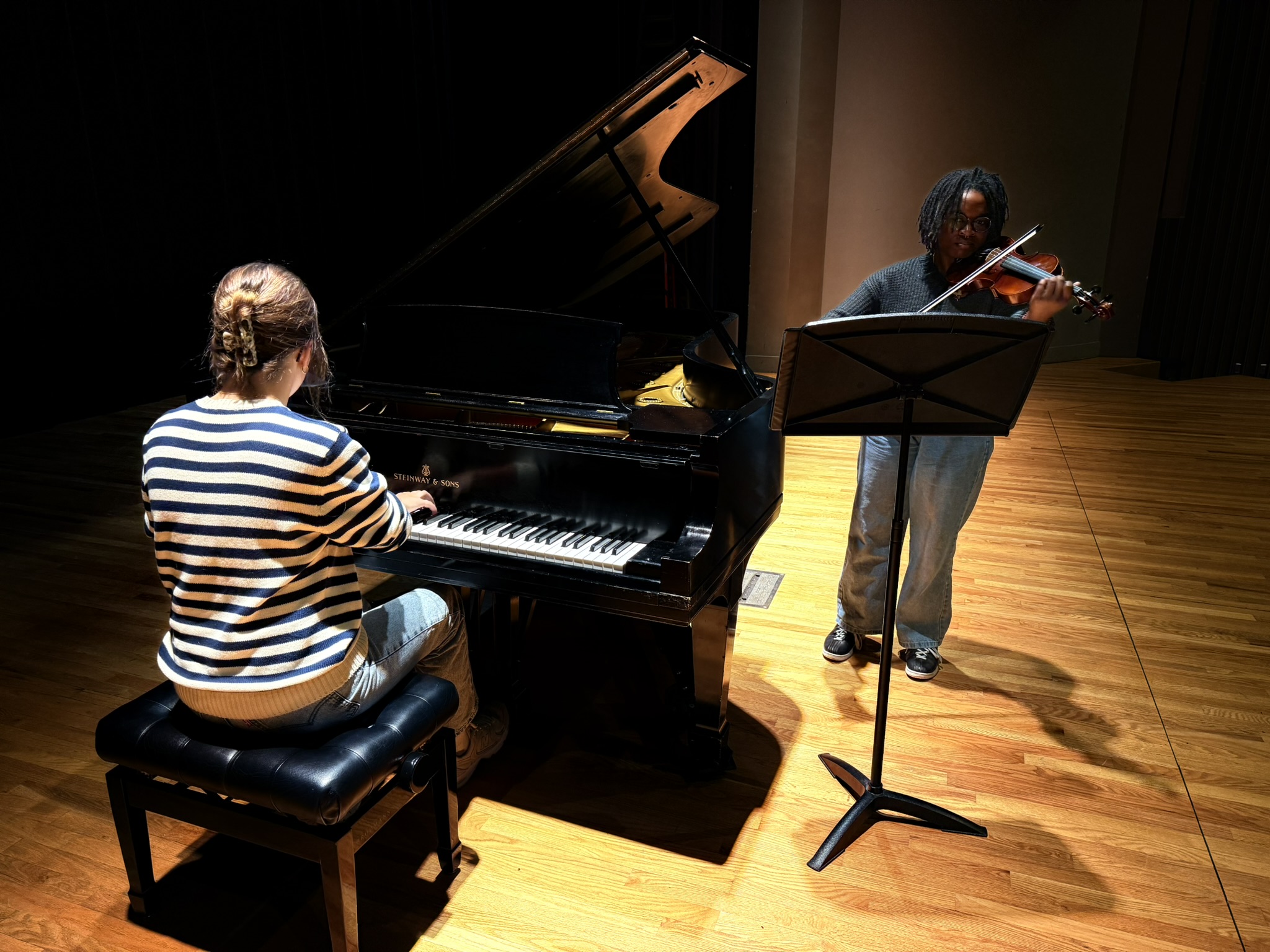 Two musicians perform on stage; one plays a grand piano while seated, wearing a striped sweater, and the other stands playing a violin, using a music stand. The stage has wooden flooring and is dimly lit.