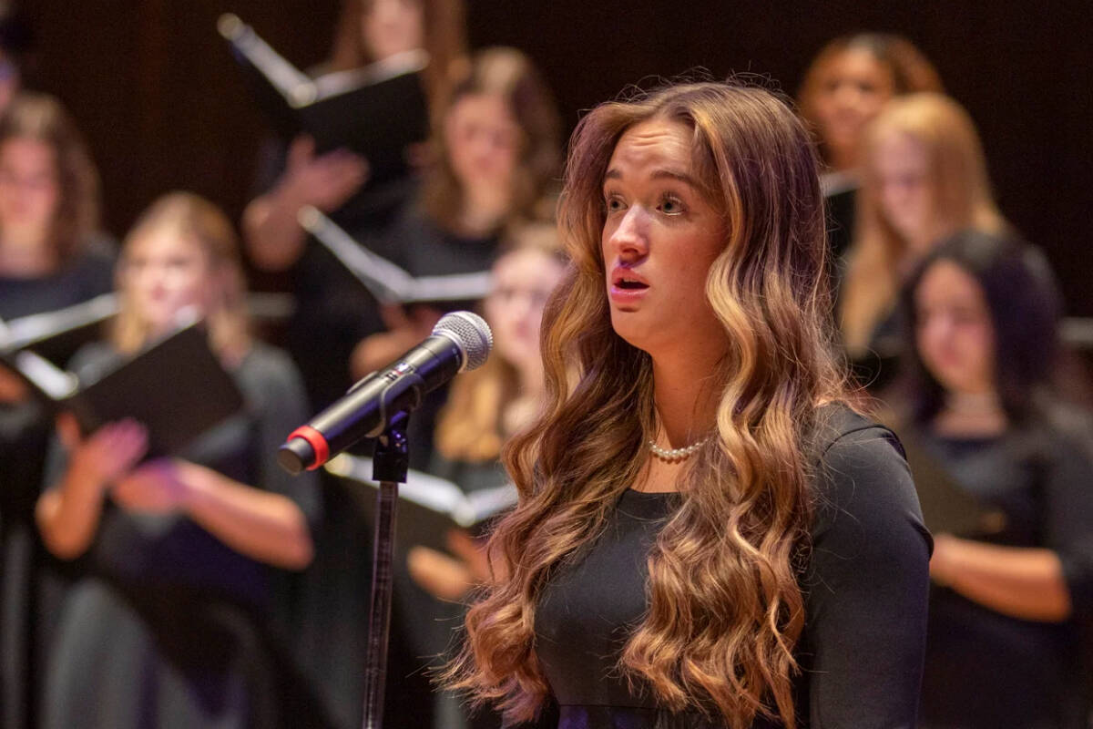 A young woman with long wavy hair stands in front of a microphone, singing. She is wearing a long, dark dress. In the background, a choir of women in similar attire is also singing, holding black folders with sheet music. The setting appears to be a concert hall.