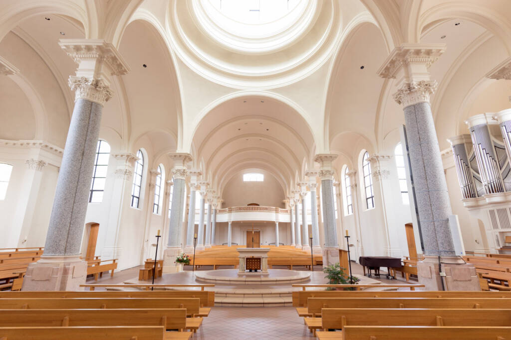 Spacious, sunlit church interior with high vaulted ceilings, tall arched windows, decorative columns, wooden pews, a central altar, and a pipe organ on the right side.