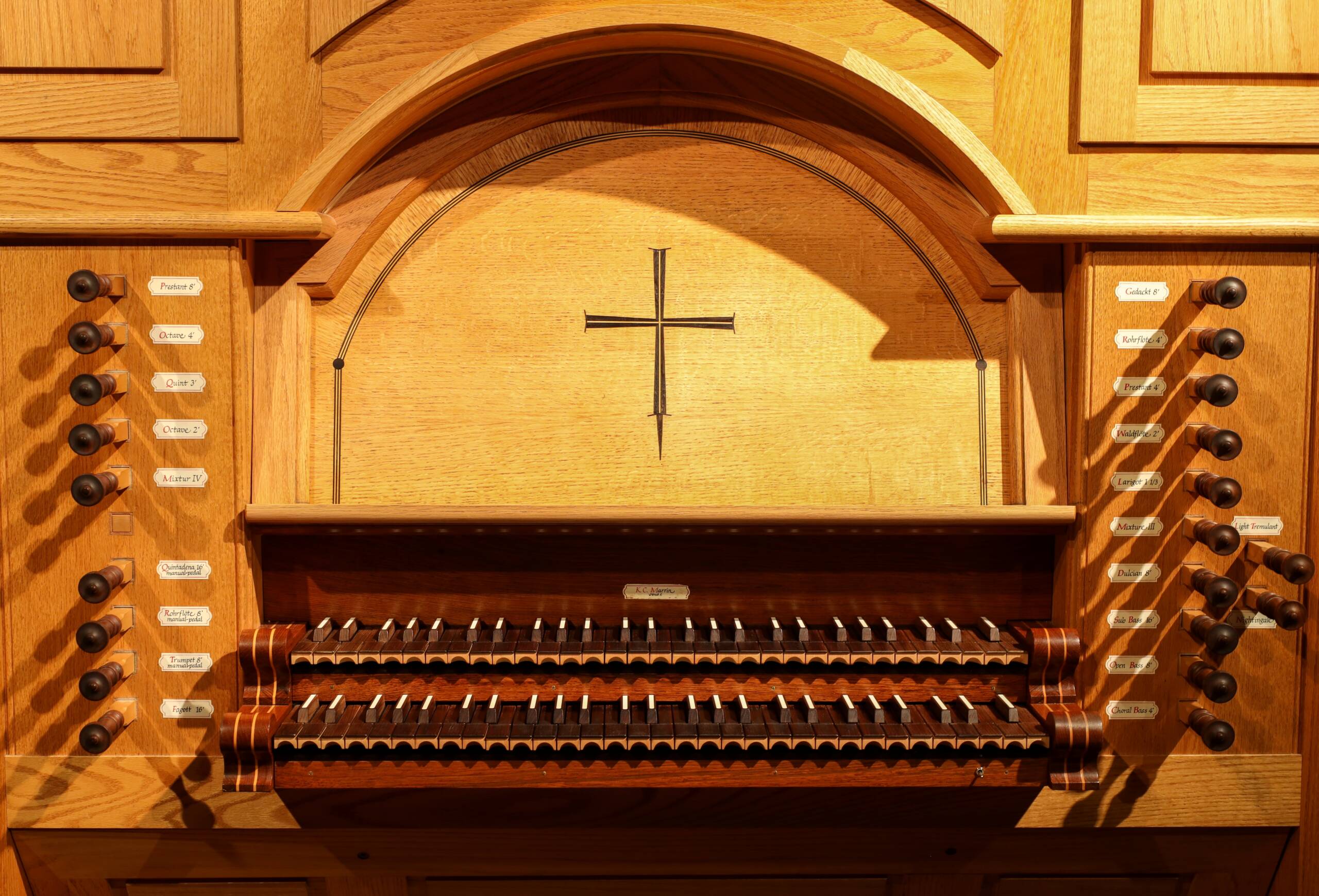 A wooden church organ console with multiple rows of keys, several labeled stops on both sides, and a metal cross above the console, all set against a paneled wooden backdrop.