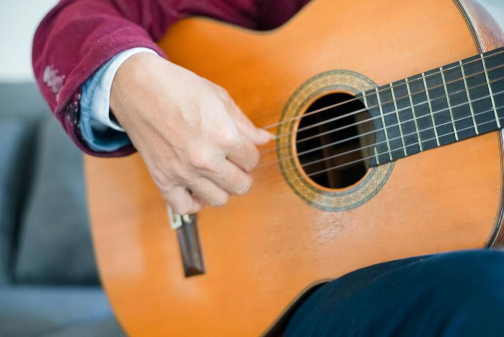 Close-up of a person’s hand strumming the strings of an acoustic guitar, wearing a maroon jacket and white shirt, with the guitar body and sound hole clearly visible.