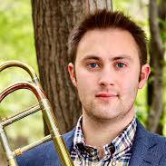 A man in a plaid shirt and blue blazer holds a brass trombone outdoors, standing in front of a tree with green foliage in the background.