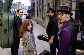 Four musicians, two men and a woman with curly hair, stand in an urban alleyway holding instrument cases, looking back toward the camera. The group wears casual, stylish clothing and hats.