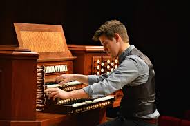 A man in a vest plays a large pipe organ with multiple keyboards and numerous control stops, concentrating as he uses both hands and adjusts the settings.