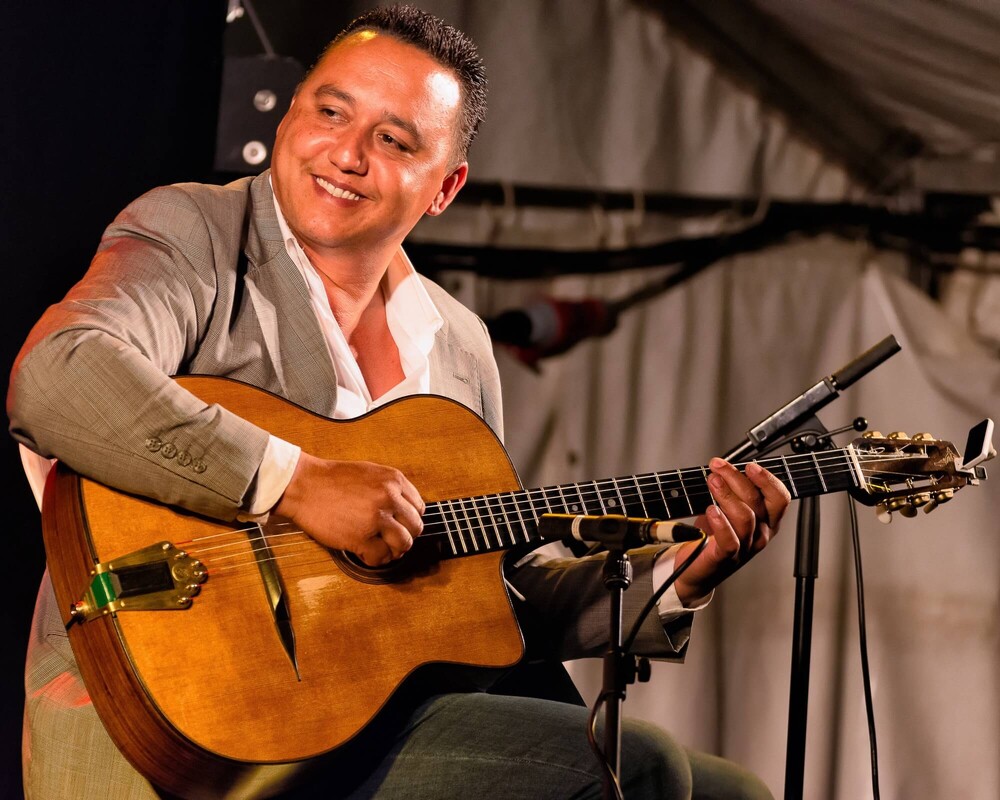 A smiling man in a gray suit plays an acoustic guitar on stage, seated under a tent, with microphones positioned in front of him.