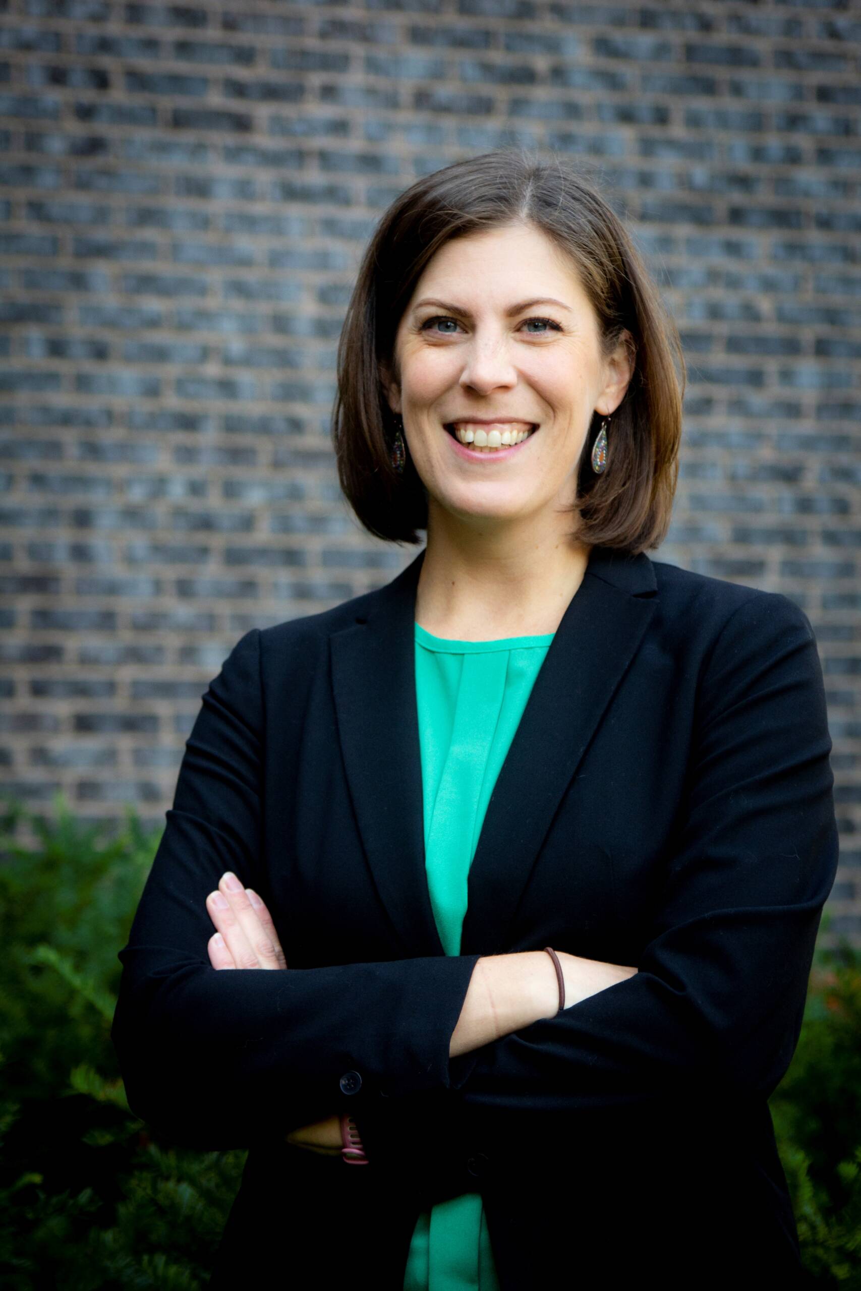 A woman with shoulder-length brown hair, wearing a black blazer over a teal blouse, stands smiling with arms crossed in front of a brick wall and greenery.