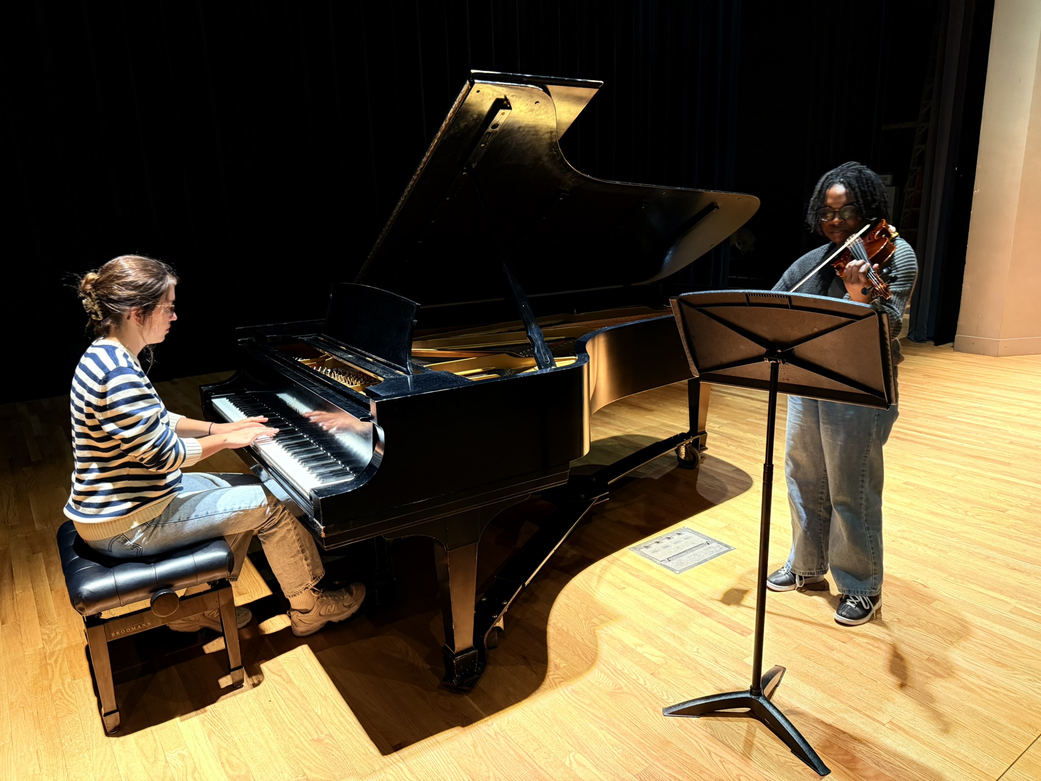 A pianist and a violinist rehearse on stage; the pianist plays a grand piano while the violinist stands next to a music stand, both focused on their performance under stage lighting.