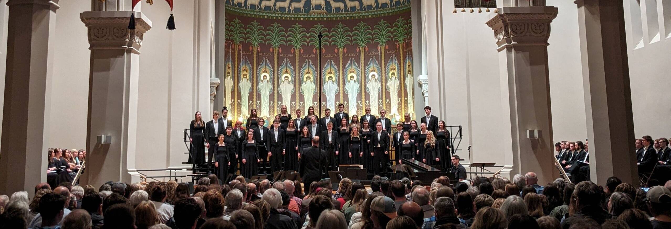 A choir performs on stage in a large church with colorful banners and a golden mosaic of Christ above the altar, as a large audience watches.