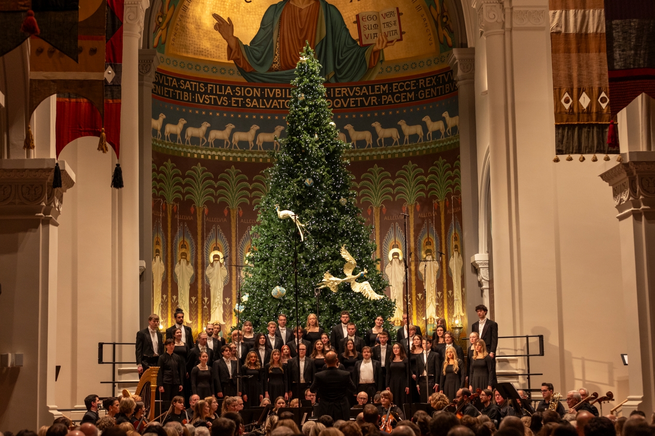 A choir performs in front of a large decorated Christmas tree inside a church with ornate architecture and a domed ceiling featuring religious artwork and golden text.