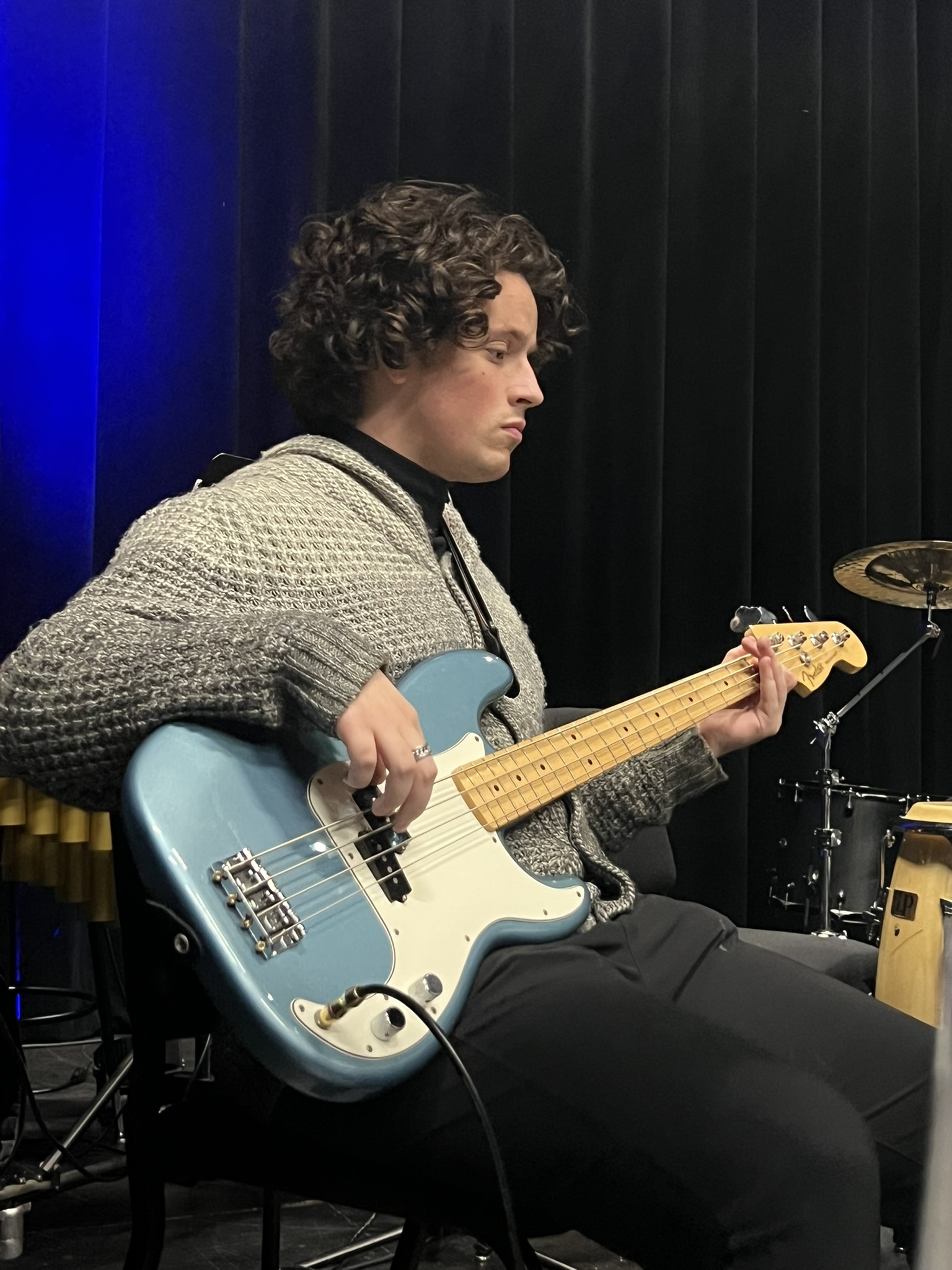 A person with curly hair sits and plays a light blue electric bass guitar, wearing a gray sweater and black pants, in front of a dark background with some musical instruments visible.
