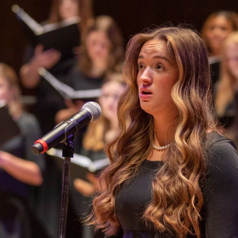 A young woman with long wavy hair stands in front of a microphone, singing. She is wearing a long, dark dress. In the background, a choir of women in similar attire is also singing, holding black folders with sheet music. The setting appears to be a concert hall.