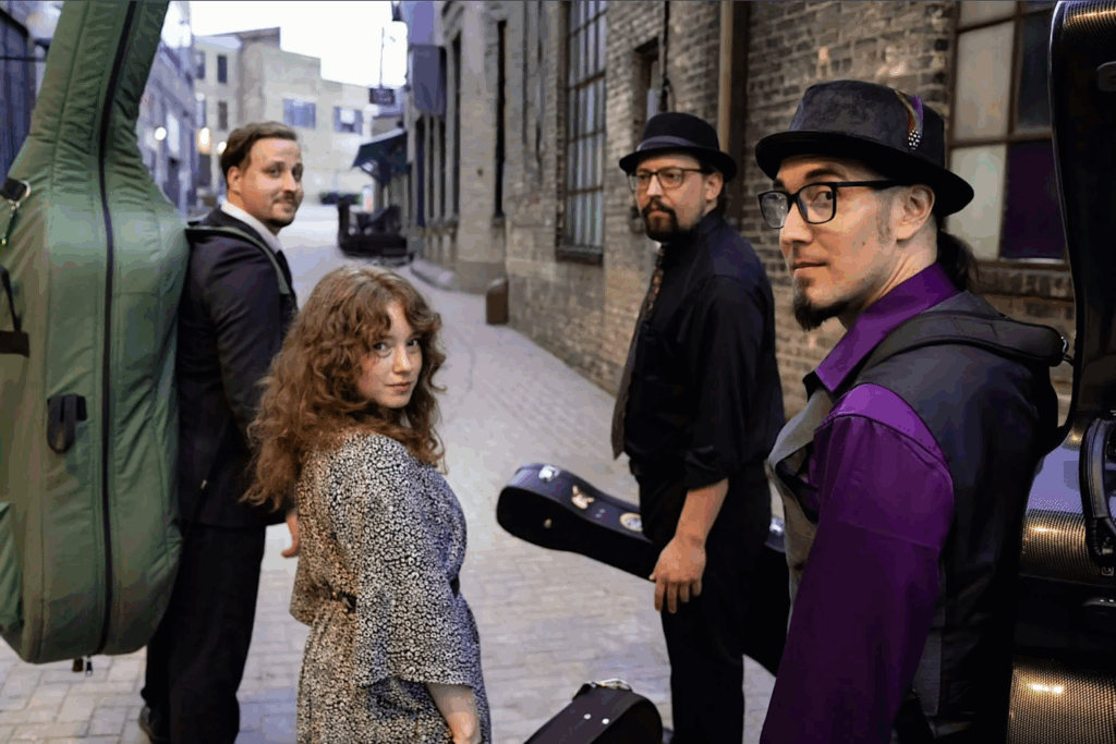 Four musicians stand in a cobblestone alleyway with their instrument cases, looking back toward the camera. The group includes two men in hats, a woman with curly hair, and a man in a suit. Brick buildings line the street.