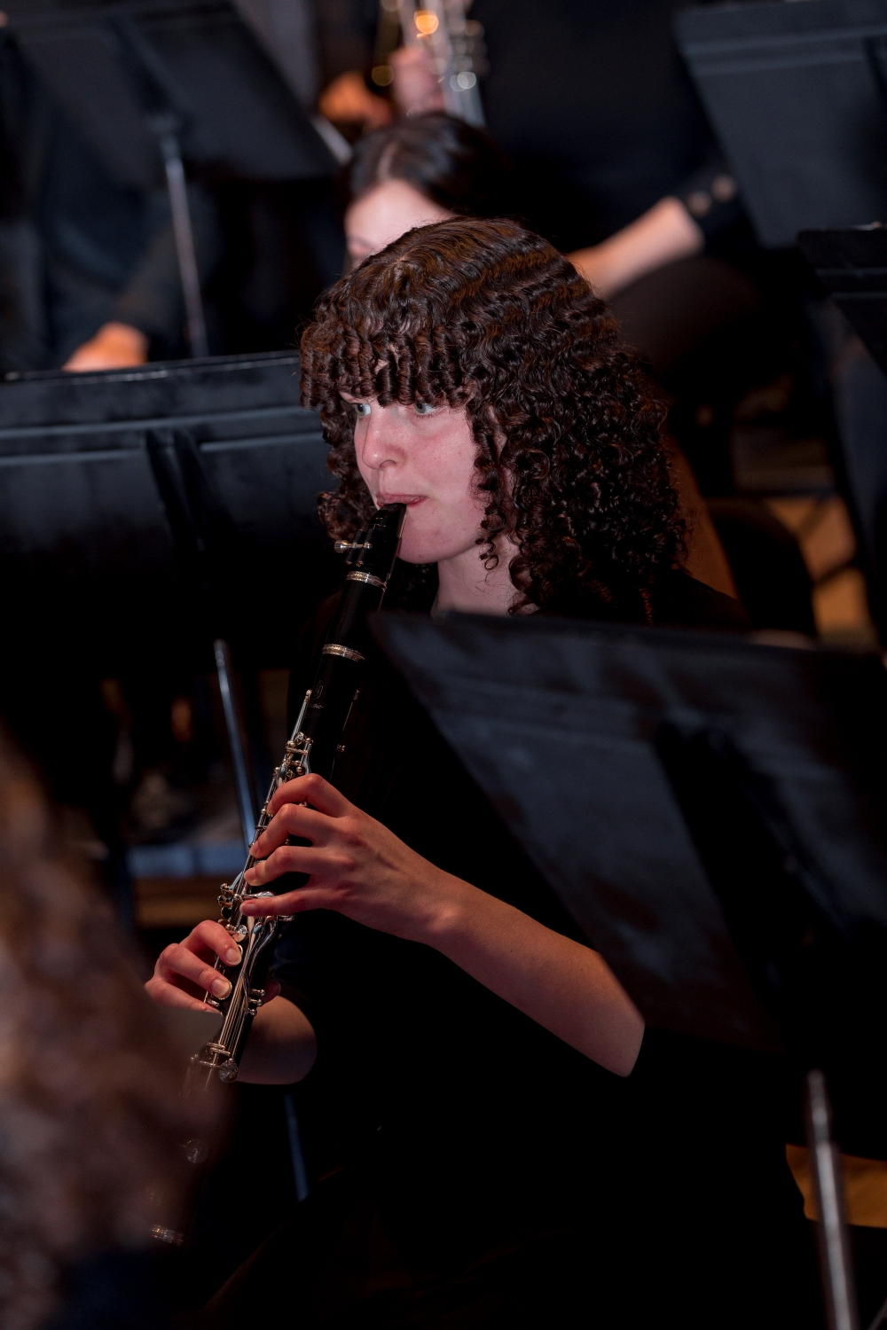 A person with curly hair plays a clarinet in an orchestra, surrounded by music stands and other musicians in a dimly lit setting.