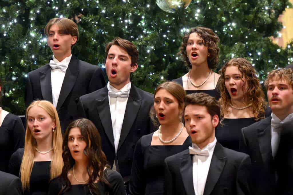 A group of young men and women in formal black attire and pearls sing in front of a decorated, lit Christmas tree. Their mouths are open mid-song, and their expressions are focused.