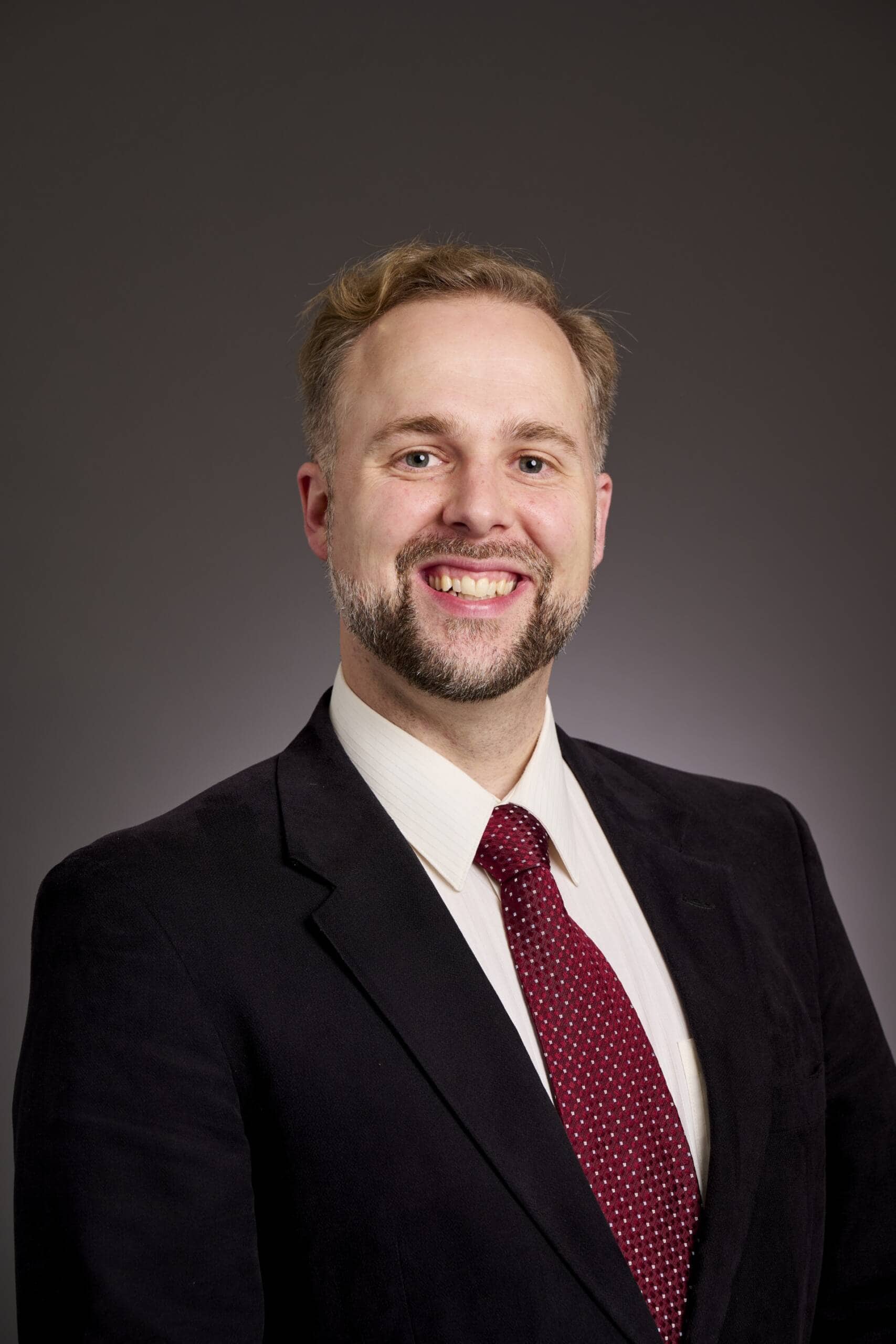 A man in a dark suit jacket, white dress shirt, and red patterned tie is smiling at the camera against a plain, dark gray background.