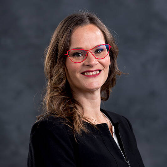 A person with long brown hair and red-framed glasses, wearing a black top, smiles and faces the camera against a dark, blurred background.