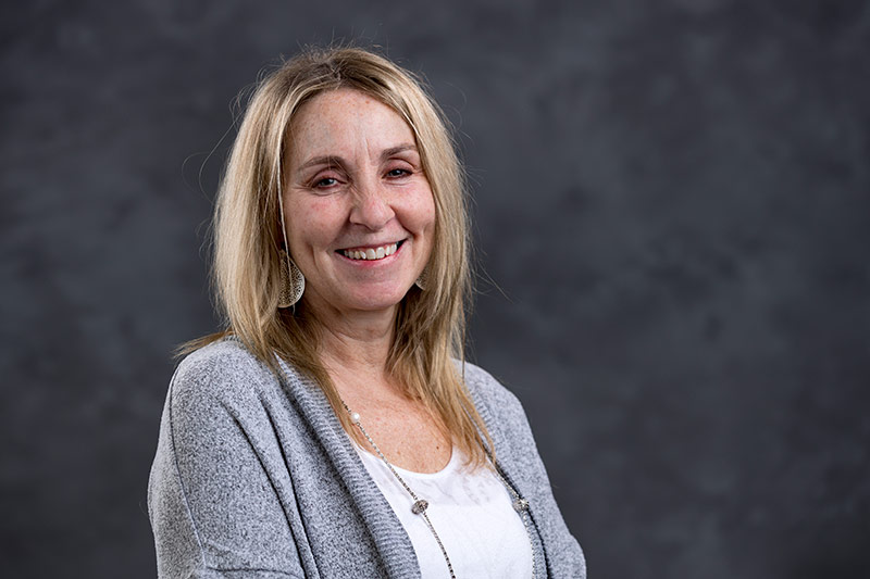 A woman with shoulder-length blonde hair smiles at the camera. She is wearing a grey cardigan over a white top. The background is solid and dark, providing a contrast to her light-colored clothing.