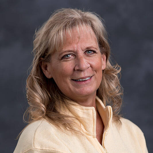 A middle-aged woman with light blond, wavy hair is smiling and wearing a light yellow zip-up fleece jacket. The background is a dark, blurred gray.