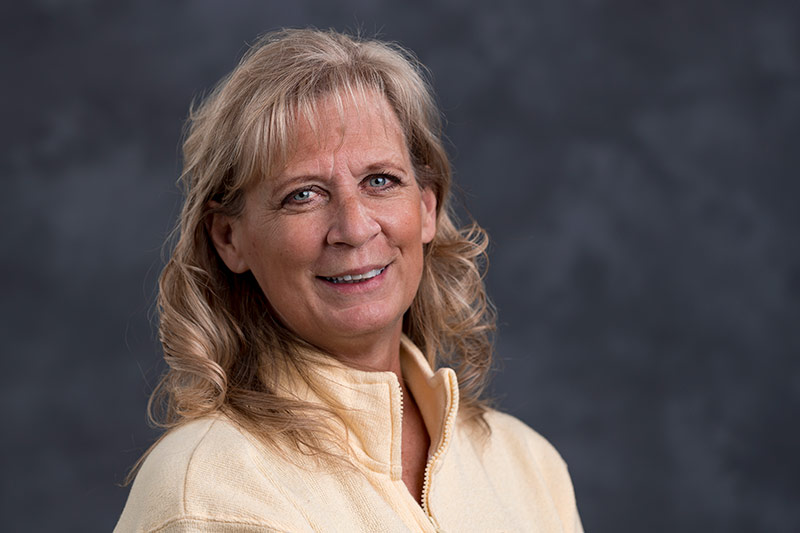 A woman with blonde, shoulder-length curly hair is smiling at the camera. She is wearing a light yellow zip-up top. The background is a plain, dark grey color.