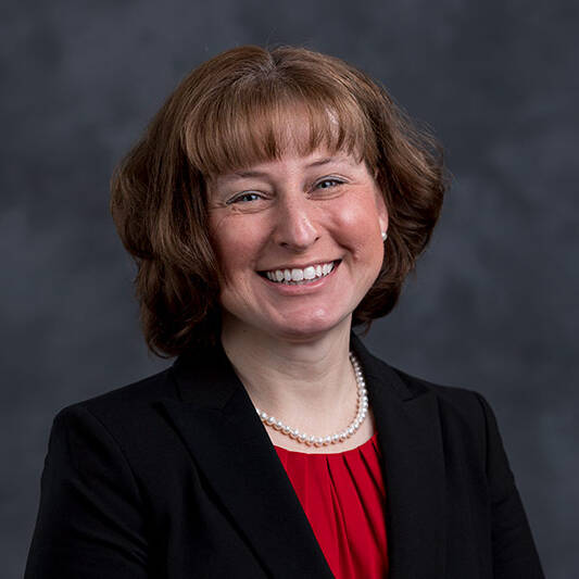 A woman with short brown hair wearing a black blazer, red top, and pearl necklace smiles in front of a dark gray, textured background.