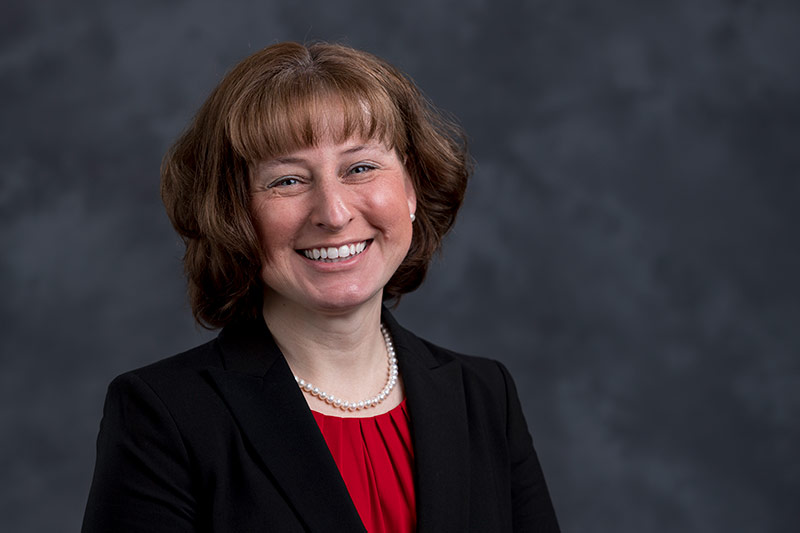 A woman with shoulder-length brown hair, wearing a black blazer over a red top, and a pearl necklace, is smiling at the camera. The background is a dark gray textured backdrop.