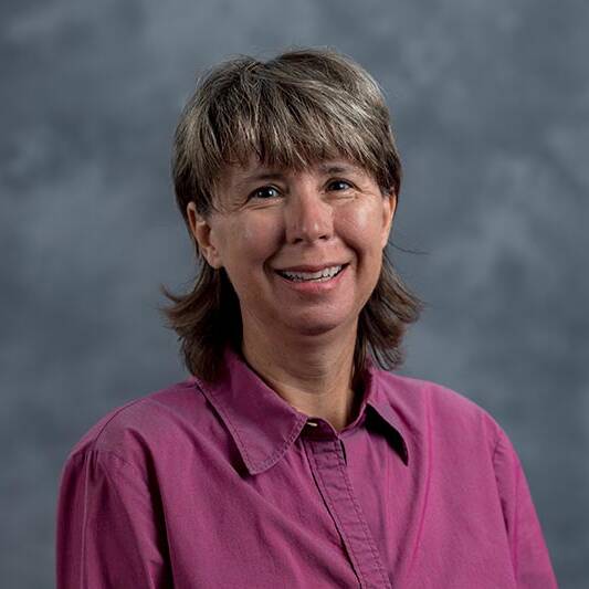 A woman with short brown hair wearing a long-sleeved magenta button-up shirt smiles in front of a gray, softly blurred background.