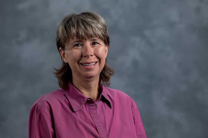 A person with short, graying hair smiles while wearing a magenta button-up shirt. The background is a neutral gray, and the lighting highlights their face.