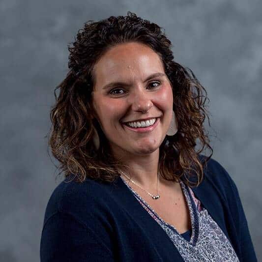 A woman with curly brown hair smiles at the camera. She is wearing a navy blue cardigan over a patterned blouse, with a simple necklace, standing in front of a gray mottled background.