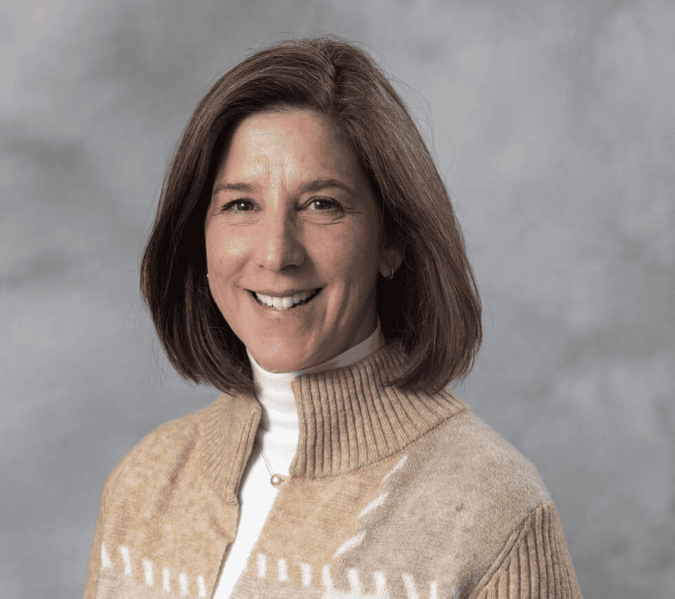 A woman with straight brown hair smiles at the camera. She is wearing a beige sweater with a white pattern and a white turtleneck. The background is a plain, light gray color.