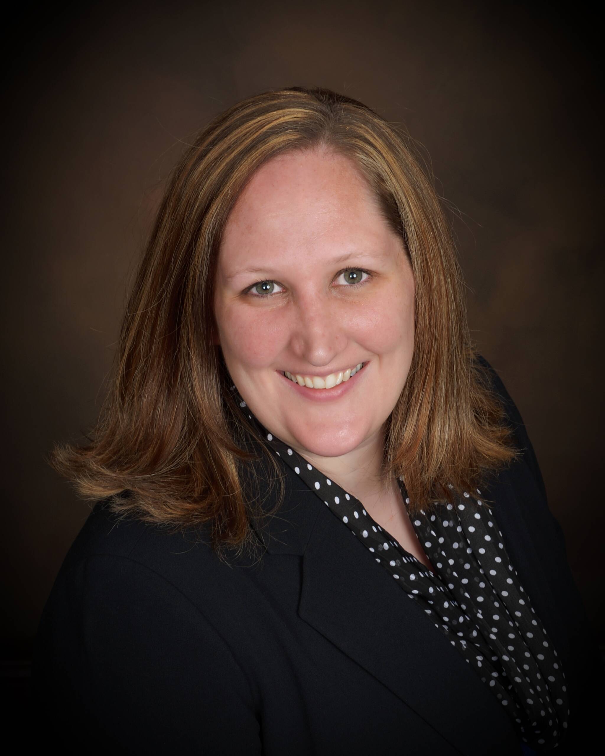 A person with shoulder-length brown hair smiles warmly at the camera. They are wearing a black jacket over a polka dot top. The background is a dark, neutral shade, adding focus to their friendly expression.