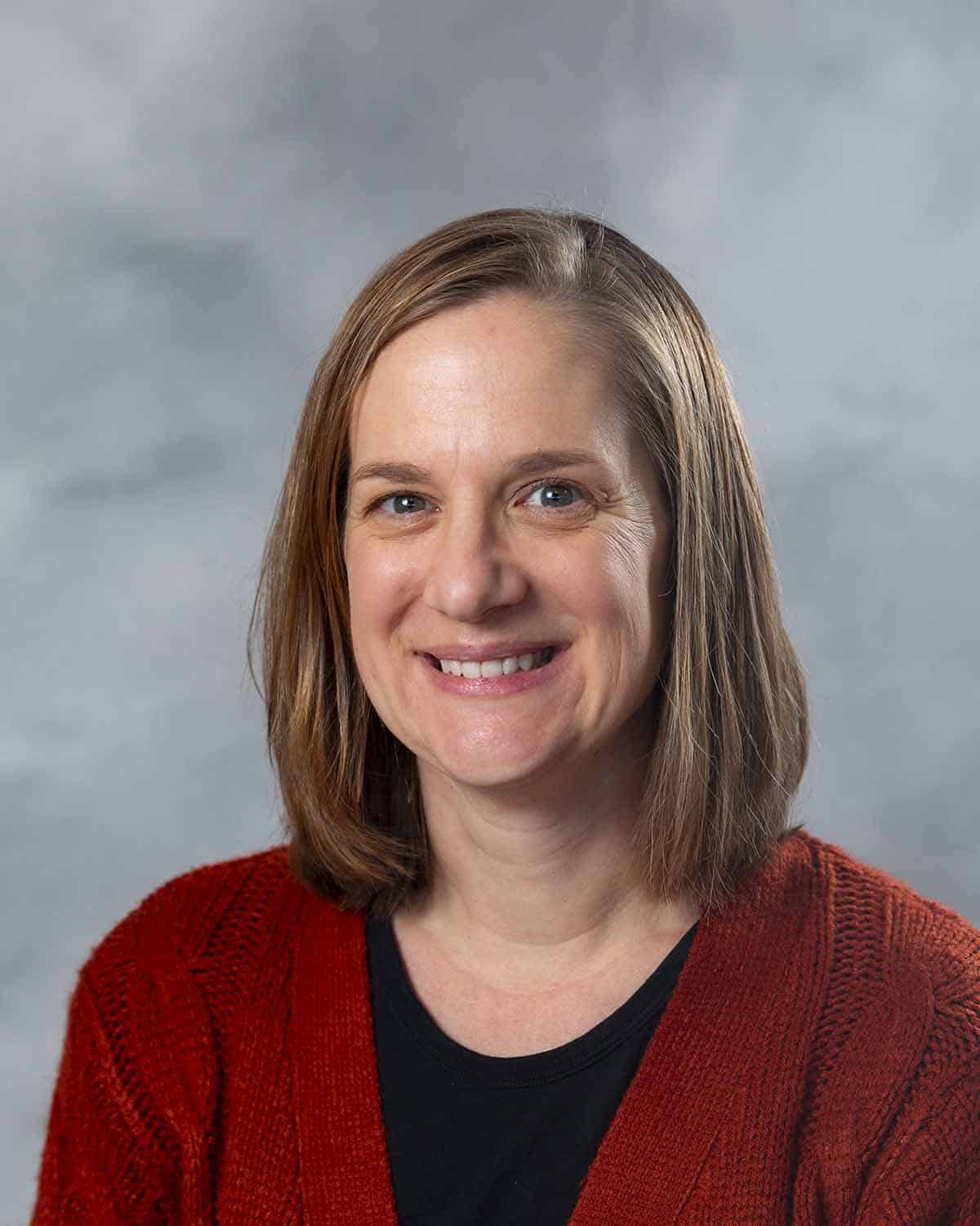 A smiling woman with straight, shoulder-length light brown hair wearing a red knit sweater over a black top, posed in front of a gray, softly blurred background.