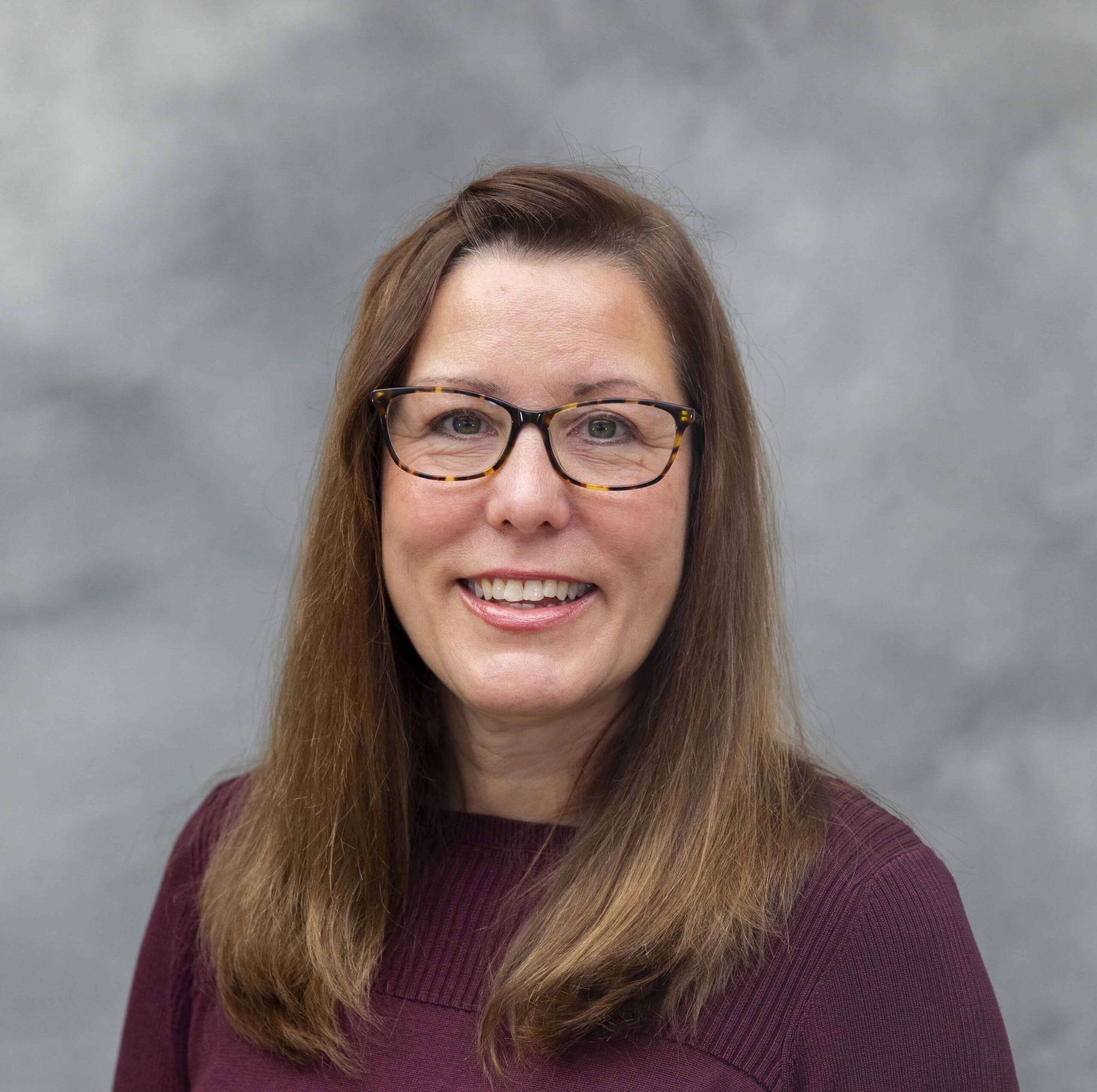 A woman with long brown hair and glasses smiles at the camera. She is wearing a burgundy sweater and is posed in front of a gray, textured background.