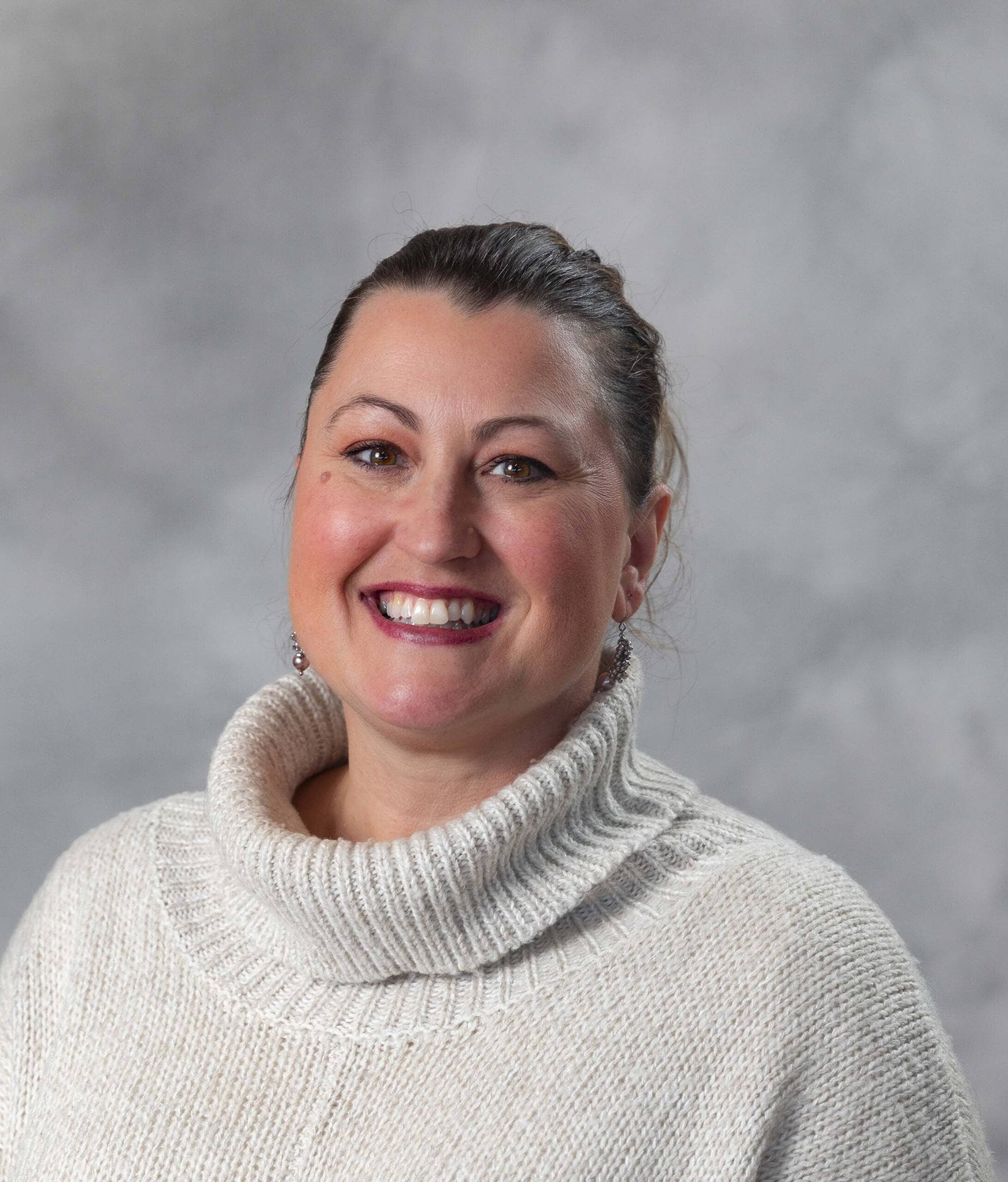 A woman with brown hair tied back, wearing a cream-colored turtleneck sweater, smiles at the camera against a light gray background.