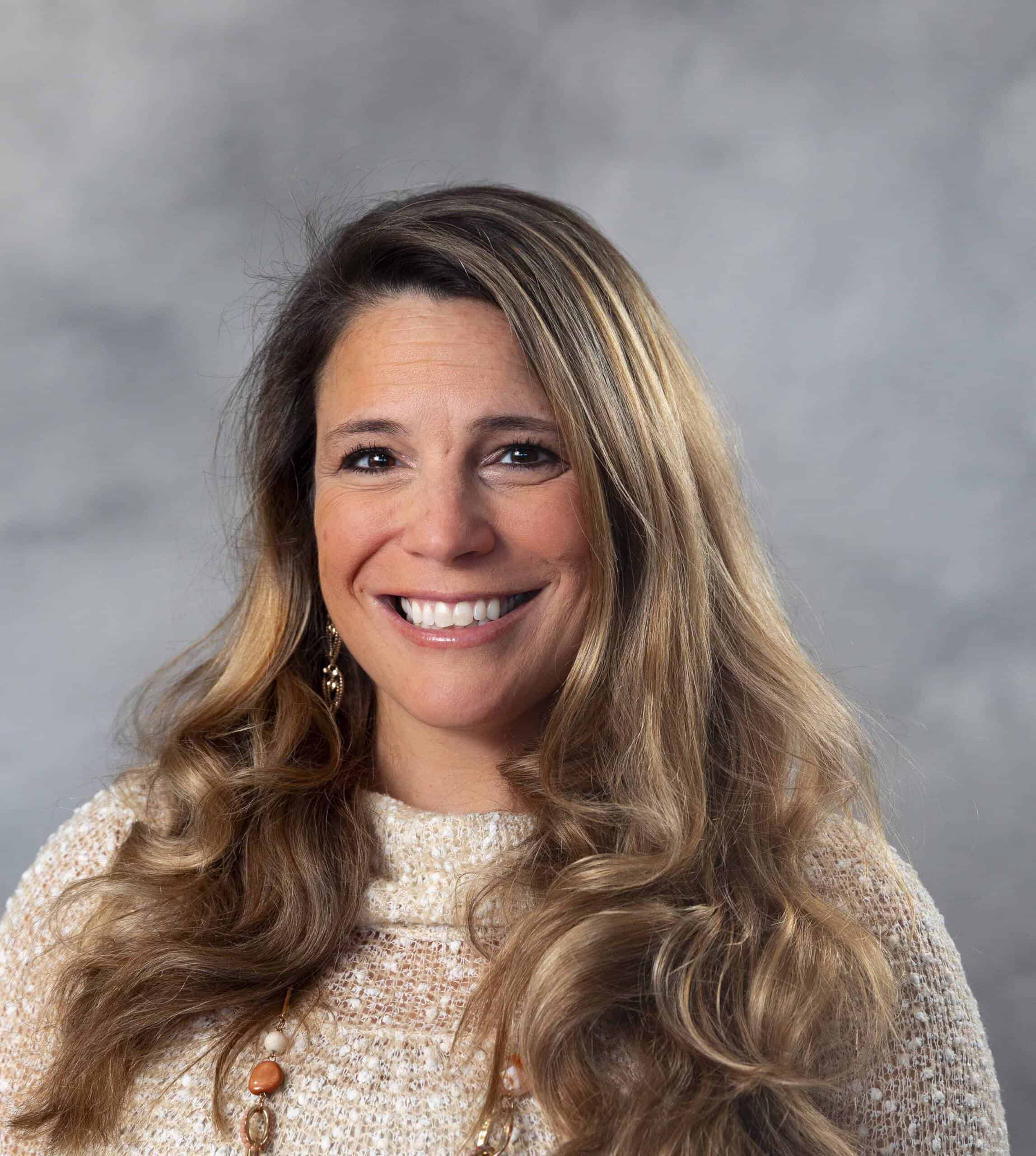 A woman with long, wavy blonde hair smiles at the camera. She is wearing a light-colored sweater and a necklace with round beads. The background is a soft, cloudy gray.