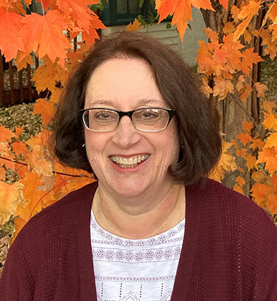 A woman with shoulder-length dark hair, wearing glasses and a maroon sweater, smiles in front of autumn leaves. The vibrant orange and yellow foliage creates a colorful backdrop.