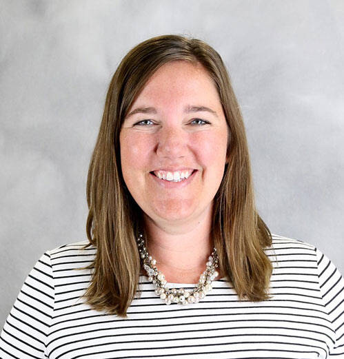 A smiling person with shoulder-length brown hair is pictured against a light gray background. They are wearing a black and white striped shirt and a pearl necklace.