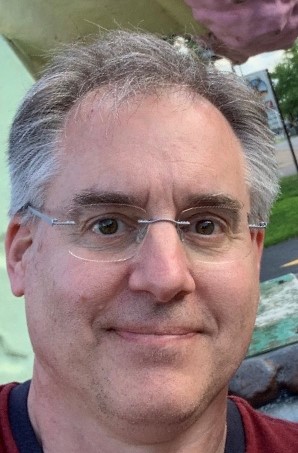 A man with short, gray hair and glasses is smiling at the camera. He is outdoors, with greenery and part of a building visible in the background. He is wearing a dark-colored shirt.