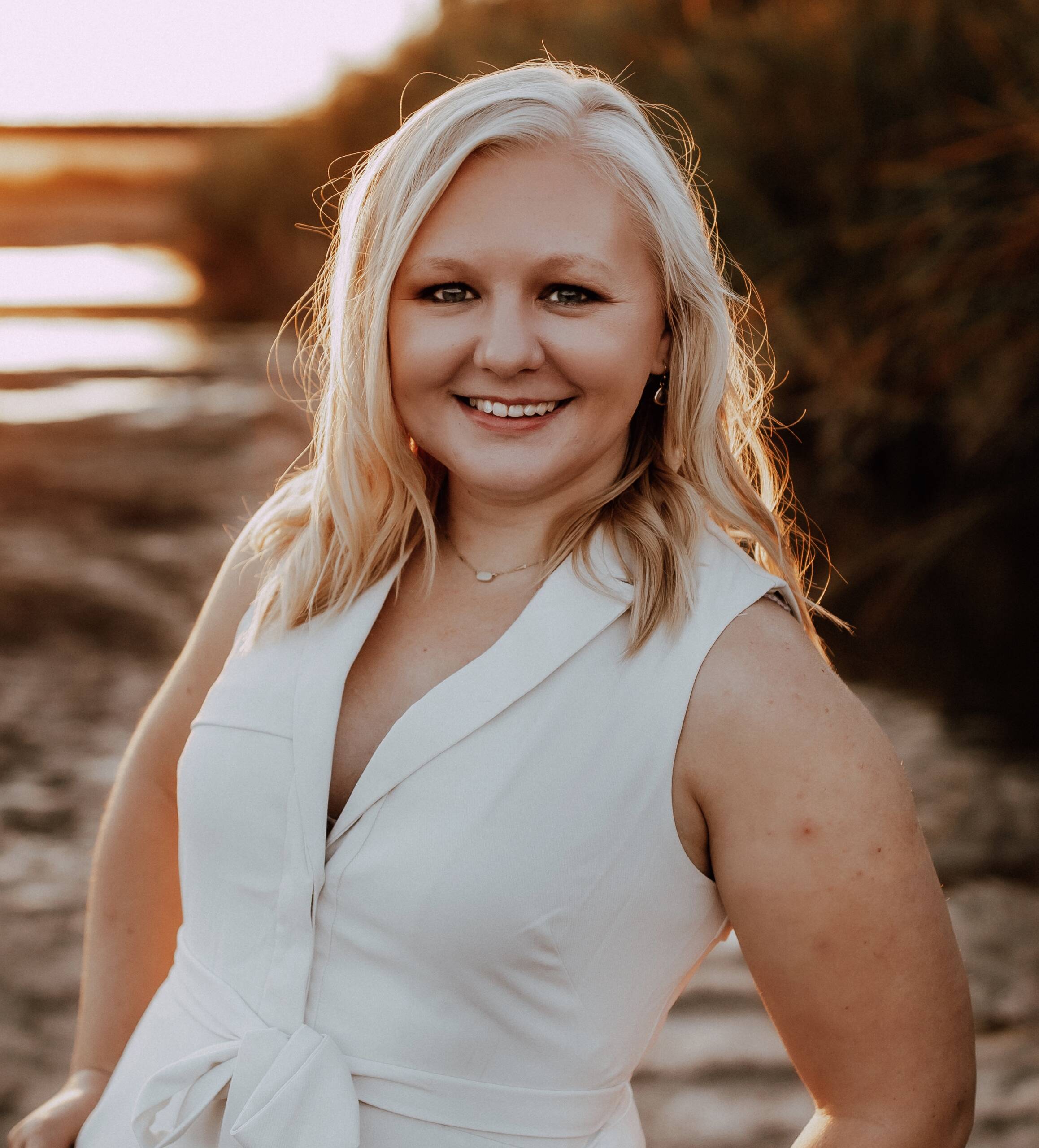 A smiling woman with long blonde hair stands outdoors near water and greenery during sunset. She is wearing a white sleeveless dress and looks directly at the camera. The warm light highlights her hair and creates a serene, golden background.