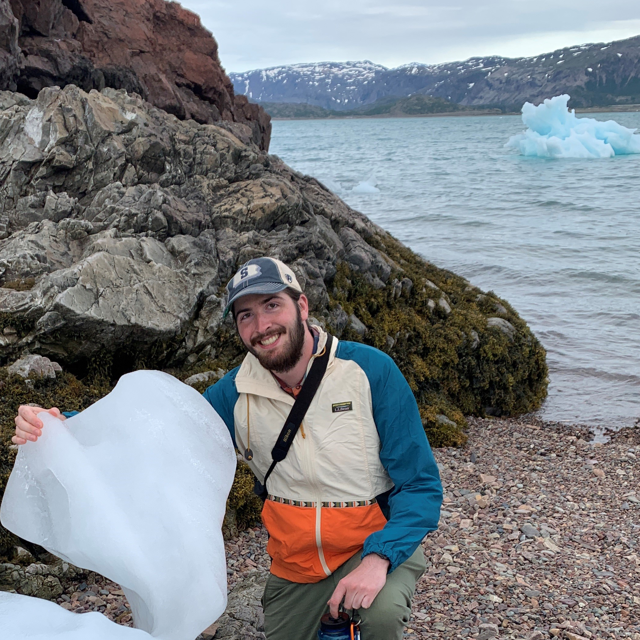 A man in an outdoor jacket and baseball cap kneels by a shoreline, holding a piece of ice in one hand. Behind him are rocky cliffs, water with floating ice chunks, and distant snow-capped mountains under a cloudy sky.