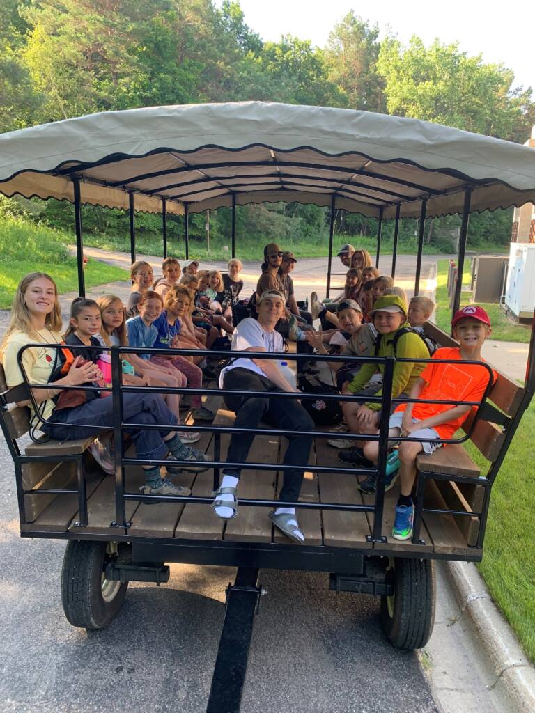 A group of people, including many children, are seated on a covered wagon, smiling at the camera. The wagon is parked on a street with greenery and trees visible in the background. The day appears sunny and bright.