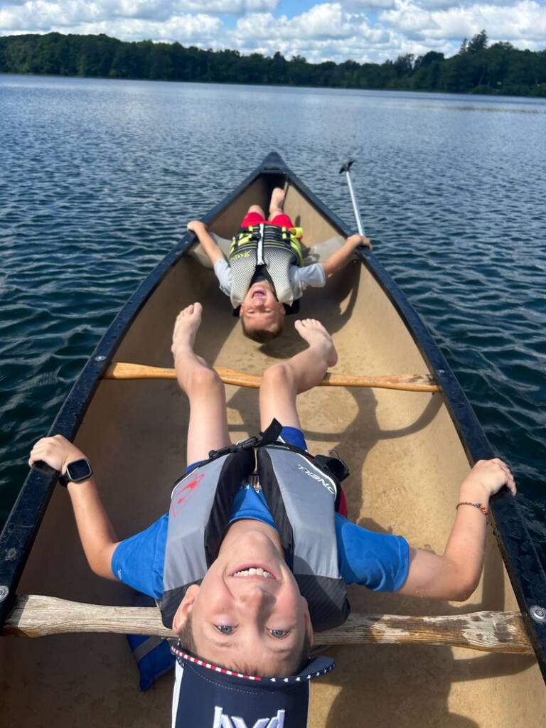 Two smiling children lie on their backs in a canoe, wearing life vests and holding paddles. They float on a calm lake under a partly cloudy sky, with a forested shoreline in the background.