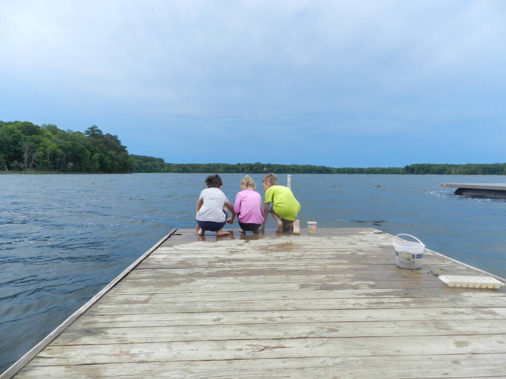 Three children sit on a wooden dock, facing a large lake under a clear sky. They're crouched at the edge, engaged in an activity. A small bucket and items are beside them. The surrounding landscape features lush green trees.