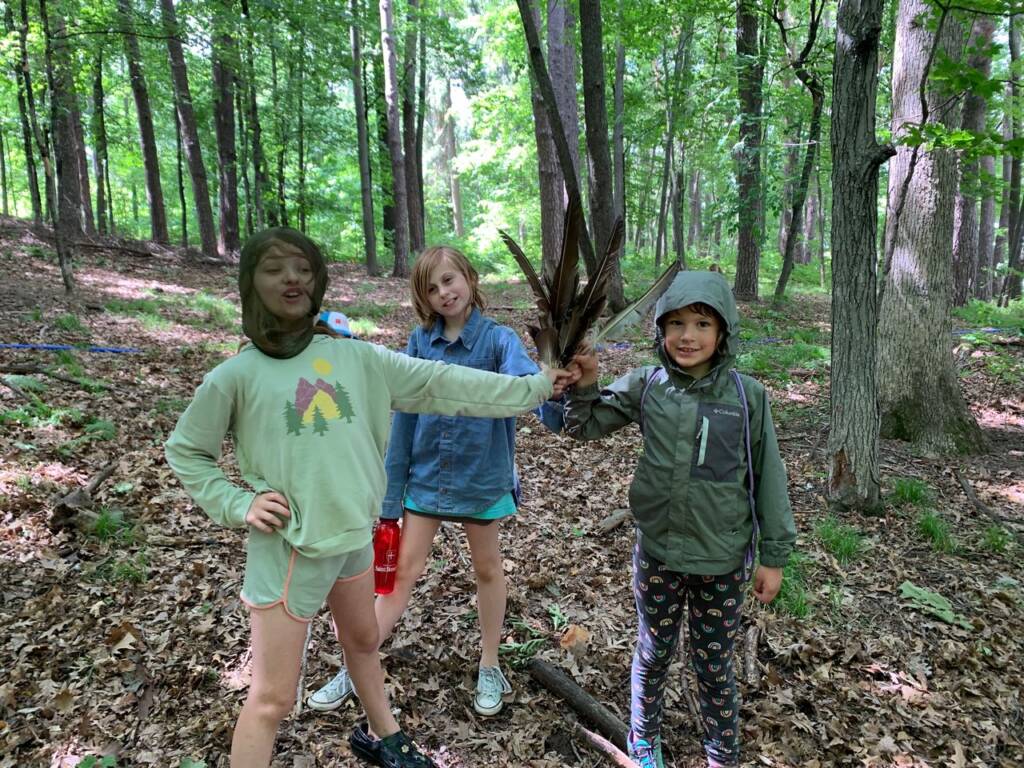 Three children stand in a forest clearing. Two wear hats and one holds twigs. The ground is covered in leaves, and trees surround them. They appear cheerful, dressed in casual outdoor attire.