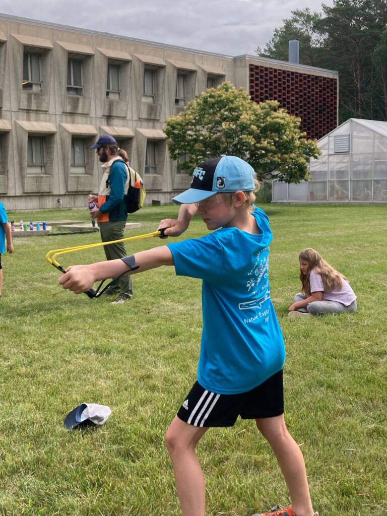 A young boy wearing a blue shirt and cap aims a slingshot on a grassy field. Nearby, a girl is crouched down, and another person with a hat stands in the background. There's a building and trees in the distance.