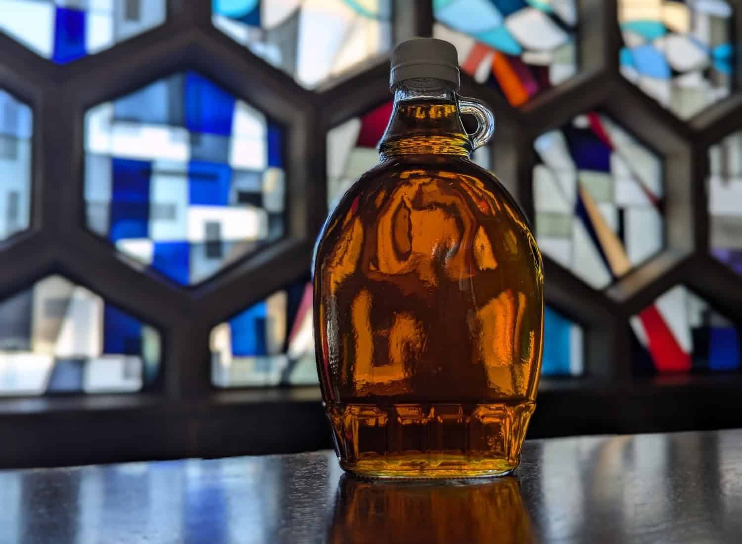 A glass bottle filled with amber liquid sits on a surface. The background features a mosaic of geometric stained glass in shades of blue, white, and black, creating an artistic and colorful backdrop.