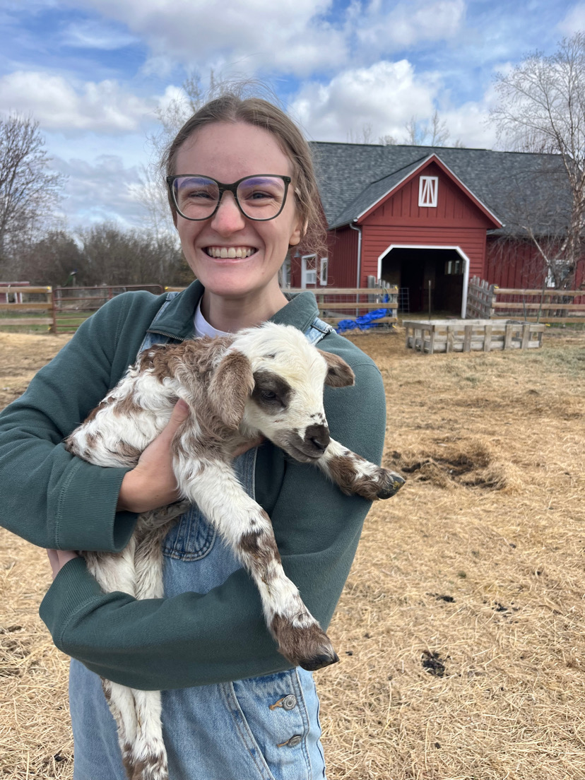 A smiling person wearing glasses and a green sweater holds a spotted baby goat outside on a farm, with a red barn and cloudy sky in the background.