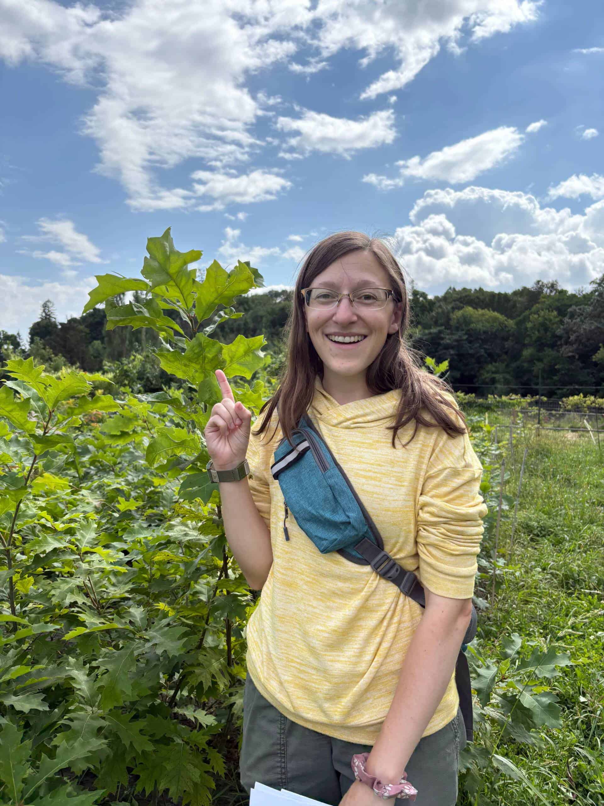 A smiling woman in a yellow hoodie stands outdoors among green plants, pointing at a leafy branch. She wears glasses, a blue crossbody bag, and holds paper. The sky is partly cloudy and trees are in the background.