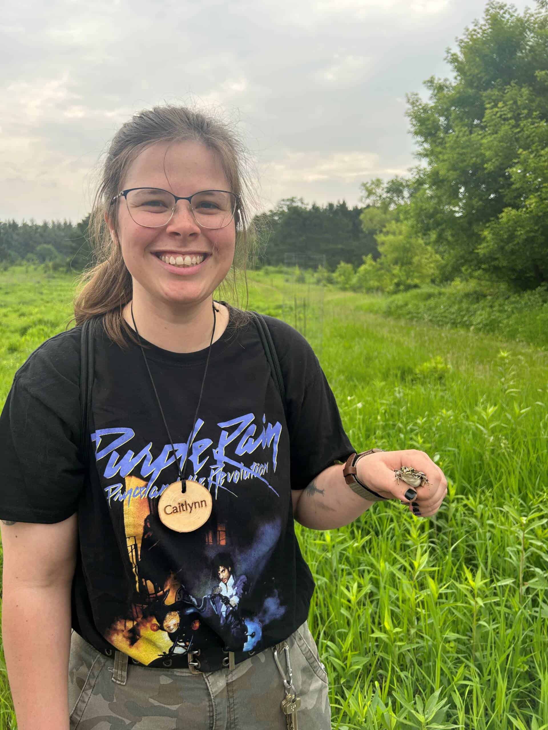 A smiling person wearing glasses and a "Purple Rain" T-shirt stands in a green field, holding a small frog and wearing a wooden name tag that says "Caitlynn." Trees and cloudy sky are in the background.