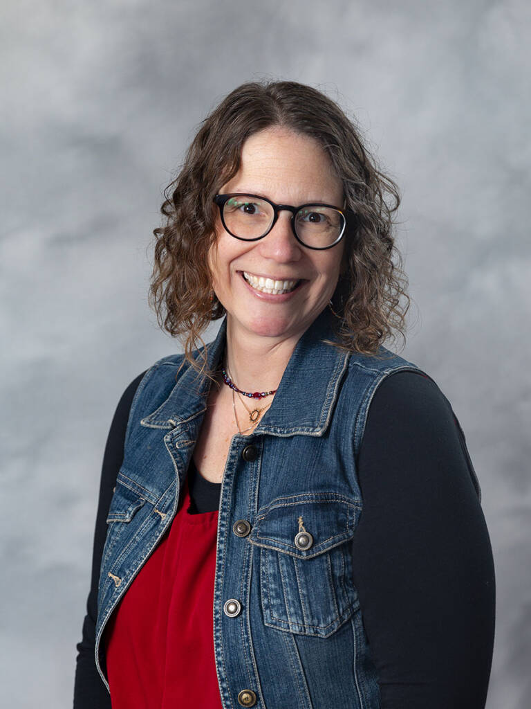 A woman with curly brown hair and glasses smiles at the camera. She is wearing a blue denim vest over a red top against a gray background.